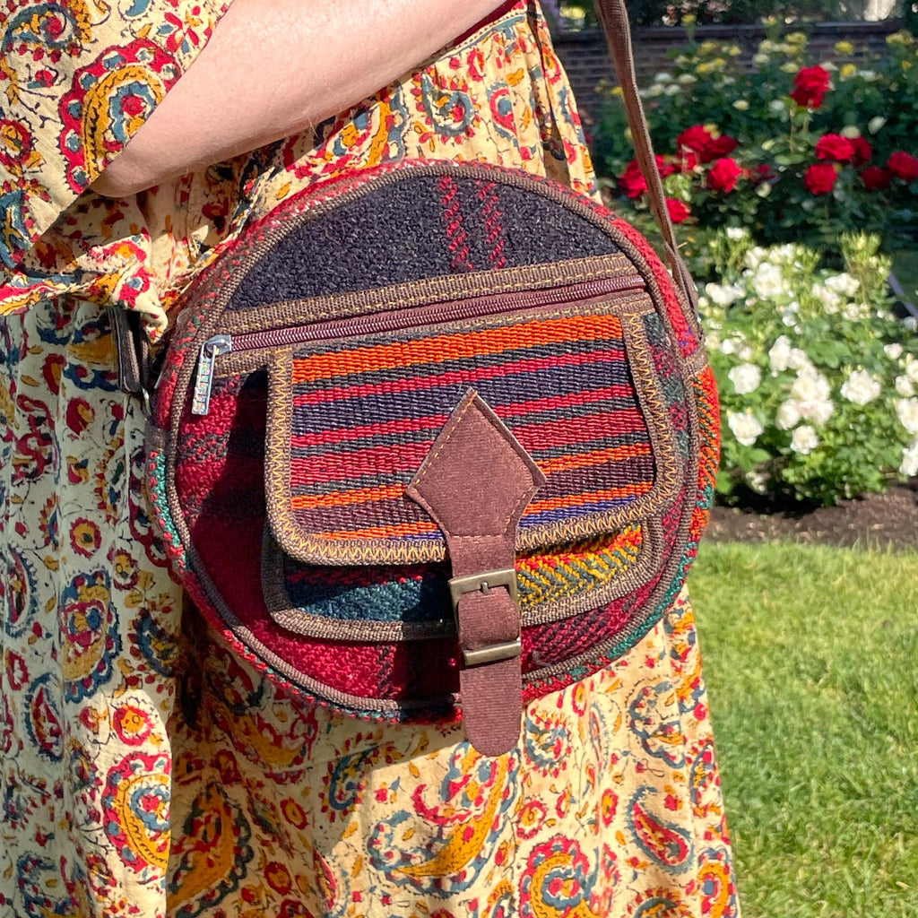 view from the front of a colorful circle bag modeled on a woman in a garden. the purse has handwoven sections of wool, trimmed with brown fabric. The bag has a long strap, front pockets, zippers, and a faux buckle over a snap closure