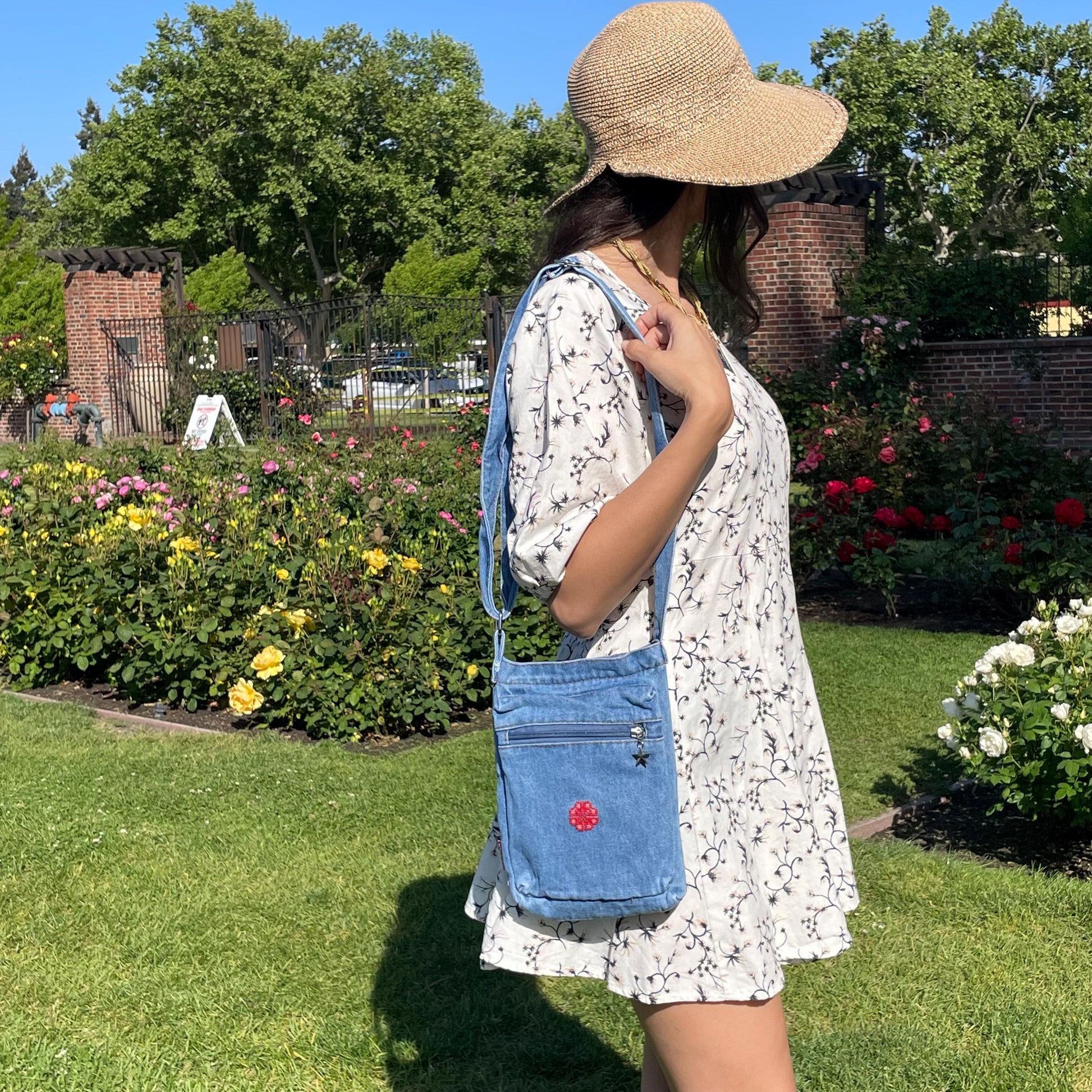 light blue denim purse with handmade tatreez embroidery flower. modeled on woman in a white dress and sun hat in a garden. view from the front