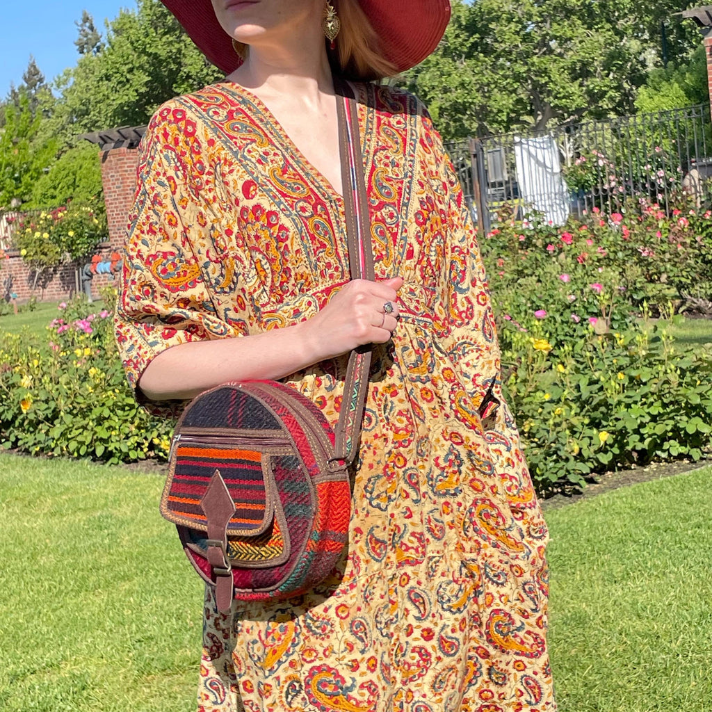 view from the side of a colorful circle bag modeled on a woman in a garden. the purse has handwoven sections of wool, trimmed with brown fabric. The bag has a long strap, front pockets, zippers, and a faux buckle over a snap closure