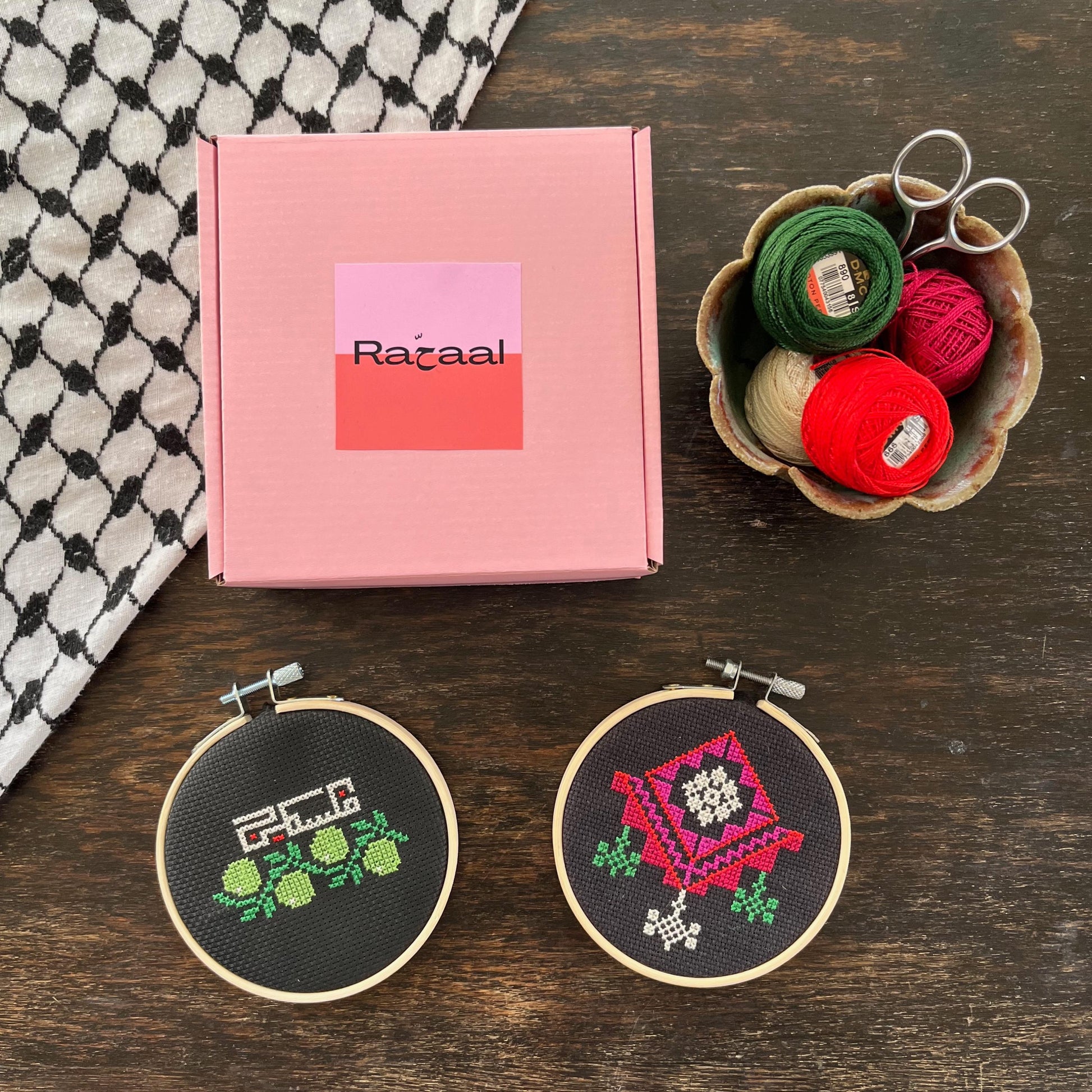 Tatreez kit with chandelier and olive branch & Palestine designs. black aida cloth, embroidery hoop, pink Rahaal tatreez kit box, embroidery scissors, and embroidery threads, view from above