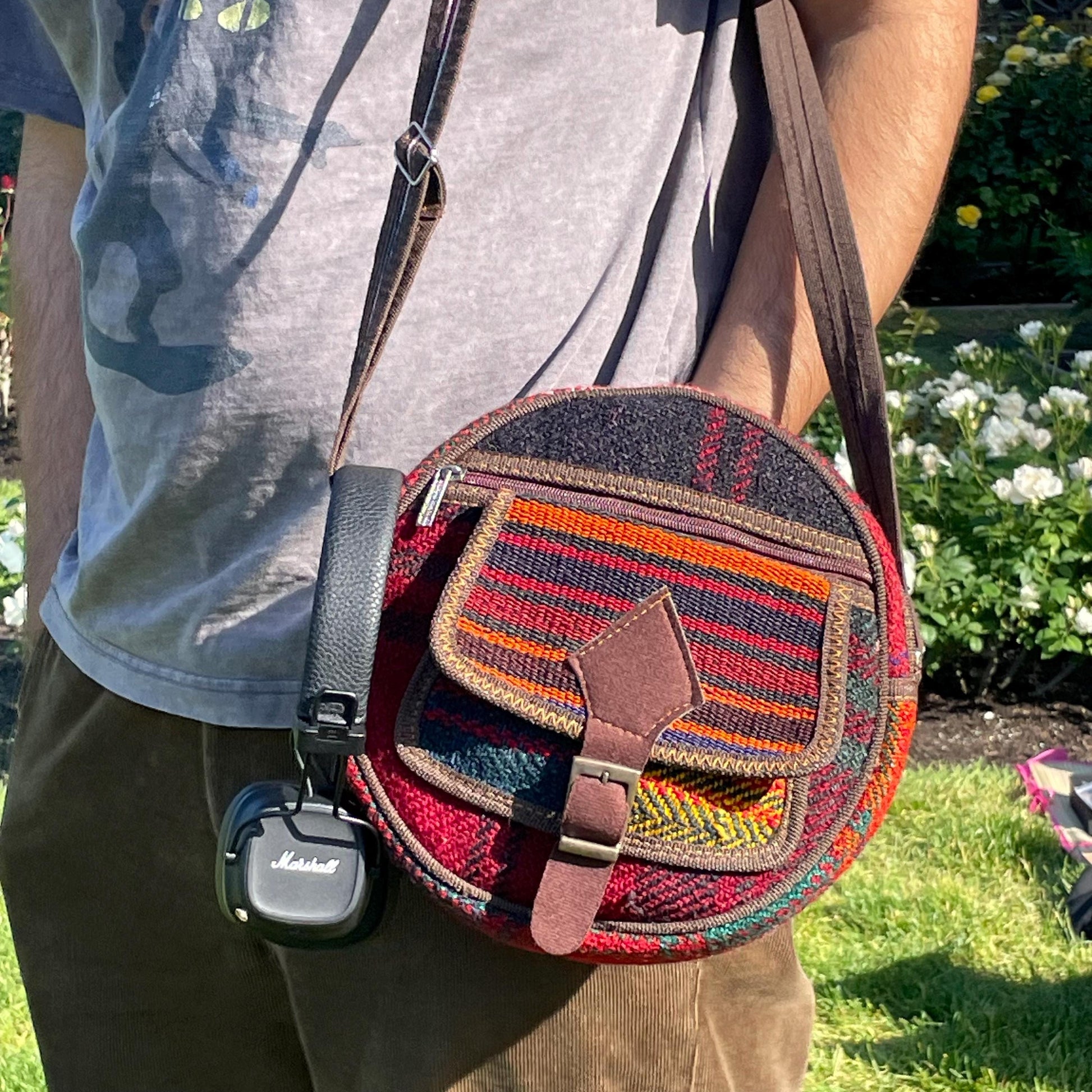 view from the front of a colorful circle bag modeled on a man in a garden. the purse has handwoven sections of wool, trimmed with brown fabric. The bag has a long strap, front pockets, zippers, and a faux buckle over a snap closure