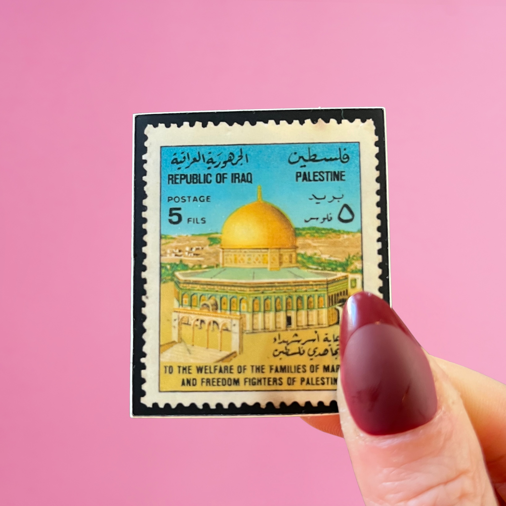 Front view of fingers holding an Iraq stamp with an image of the dome of the rock in jerusalem. Against a pink background