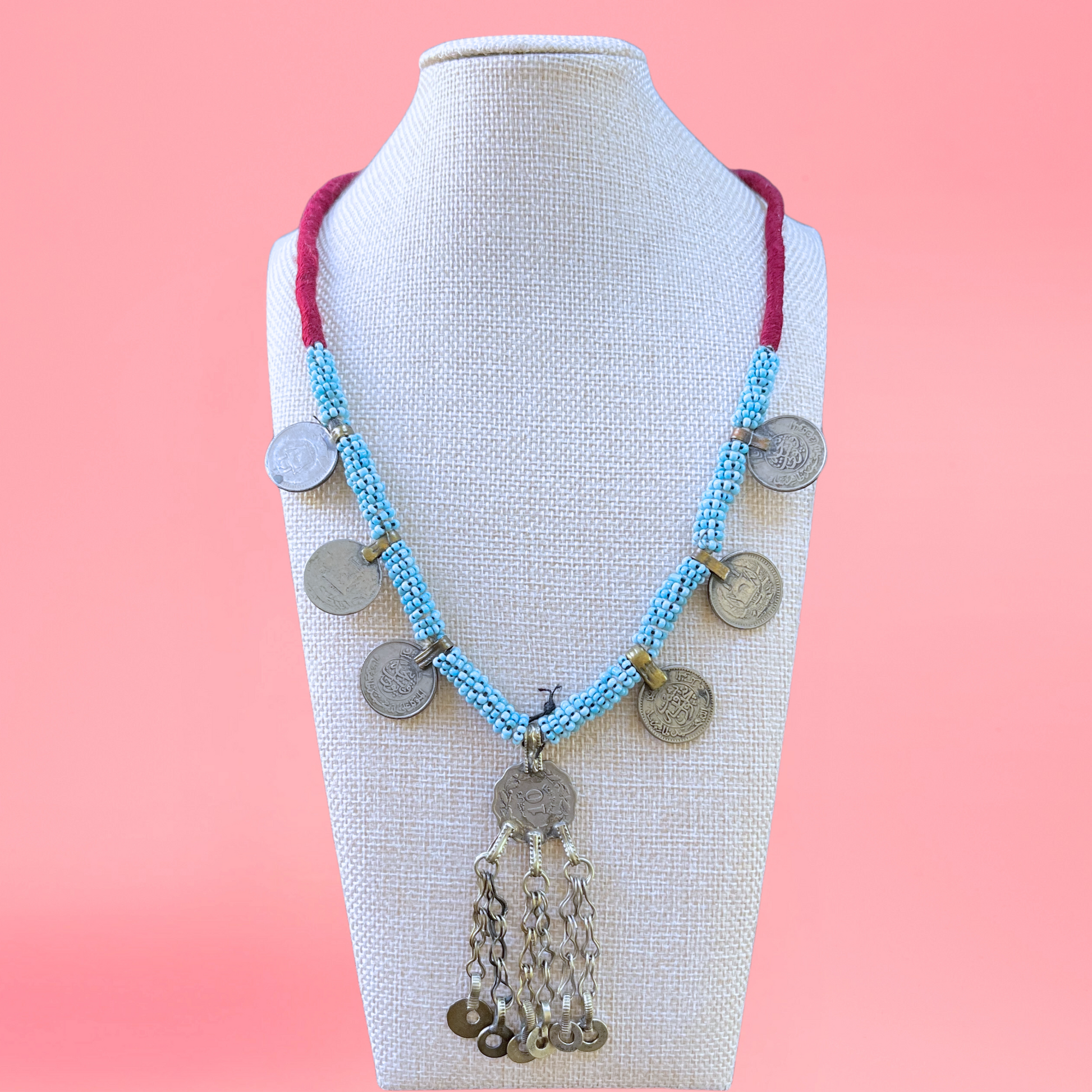 necklace modeled on a beige bust with a pink background. Necklace features persian coins, a silver pendant, and light blue beads. view from the front