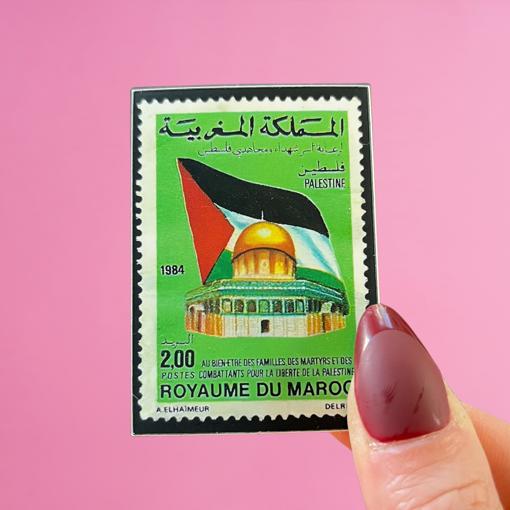 Front view of fingers holding a Morocco stamp with an image of the dome of the rock in jerusalem with a palestinian flag. Against a pink background