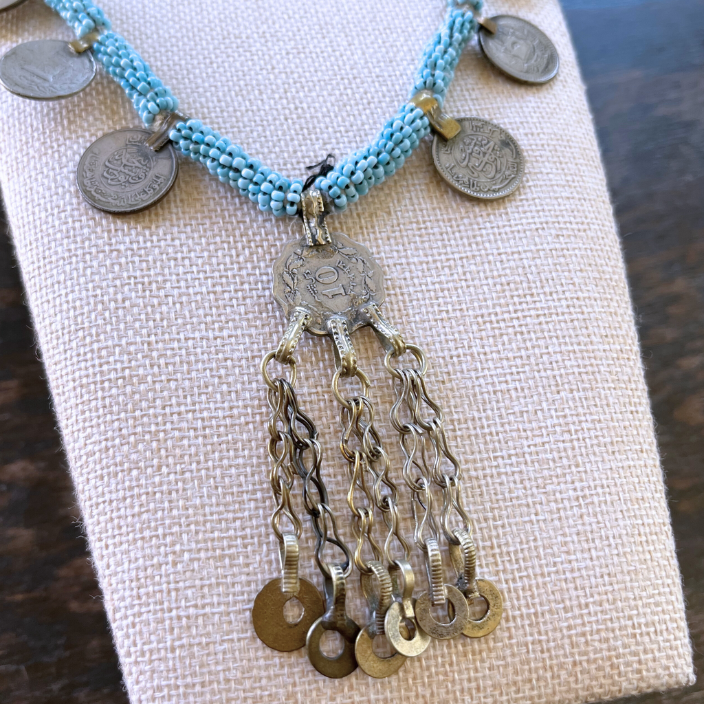 close-up view of necklace modeled on a beige bust with a pink background. a silver pendant with silver chains, and light blue beads. view from above