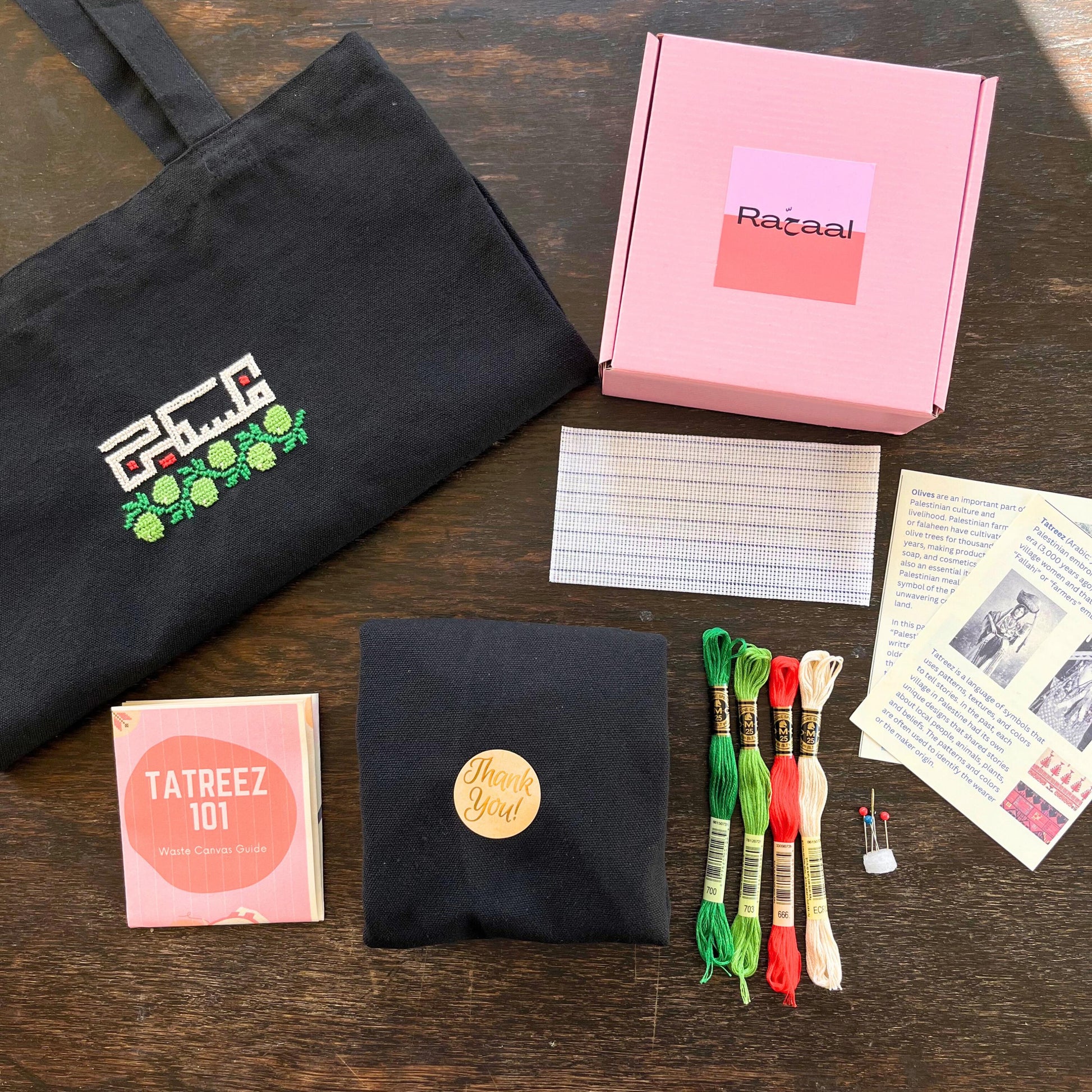 Tatreez tote bag kit with materials laid out, including black tote bag, tatreez instructions, olive and Palestine tatreez pattern, embroidery threads, embroidery needle and pins, and waste canvas. view from above