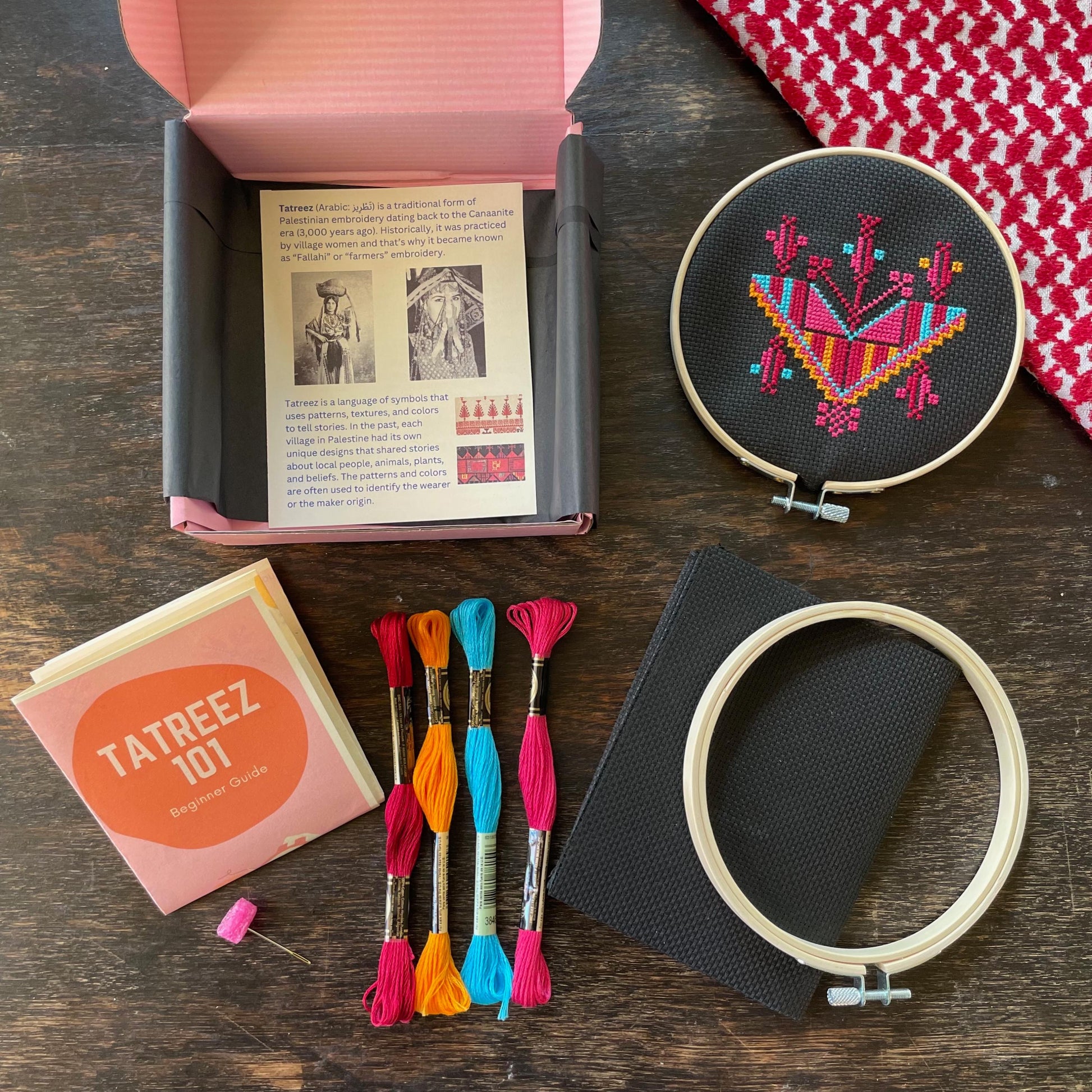 Tatreez kit with materials laid out, including black aida cloth, tatreez instructions, Gaza necklace tatreez pattern, embroidery threads, embroidery needle, and embroidery hoop. view from above