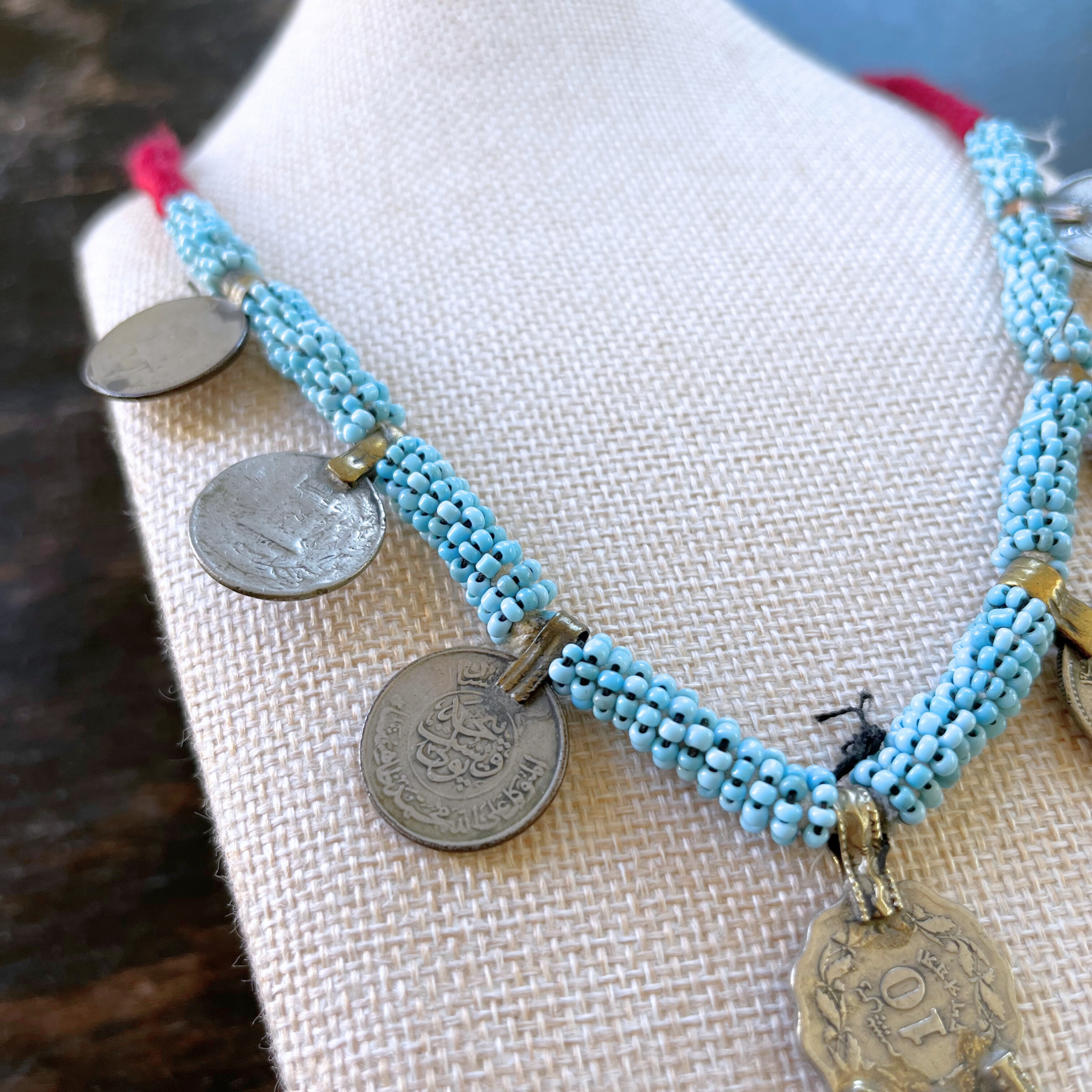 close-up view from above of a necklace modeled on a beige bust. Necklace features vintage persian coins, a silver pendant, and light blue beads.