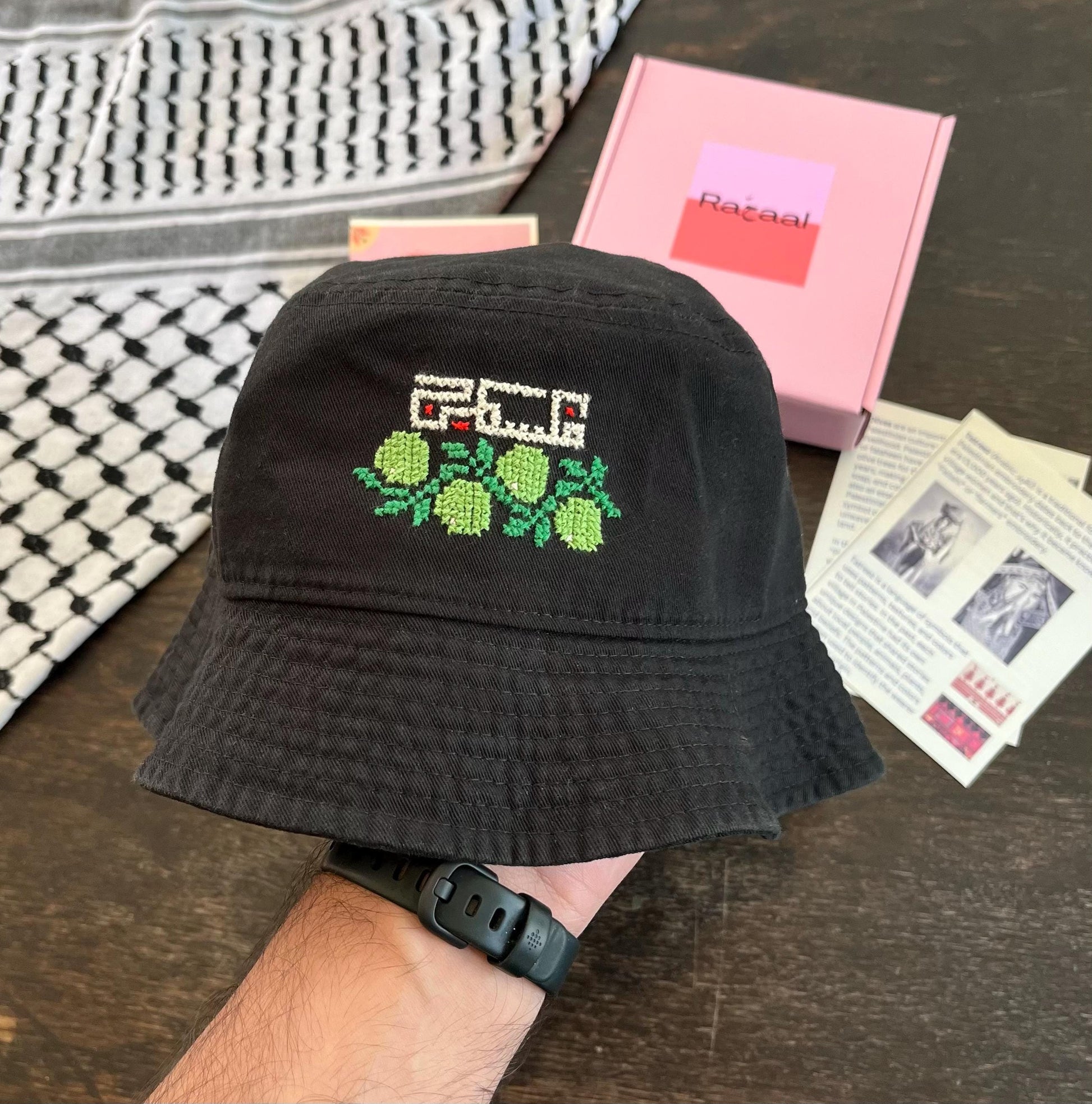 Tatreez bucket hat kit with olive branch and Palestine design. black bucket hat, pink Rahaal tatreez kit box, view from the front