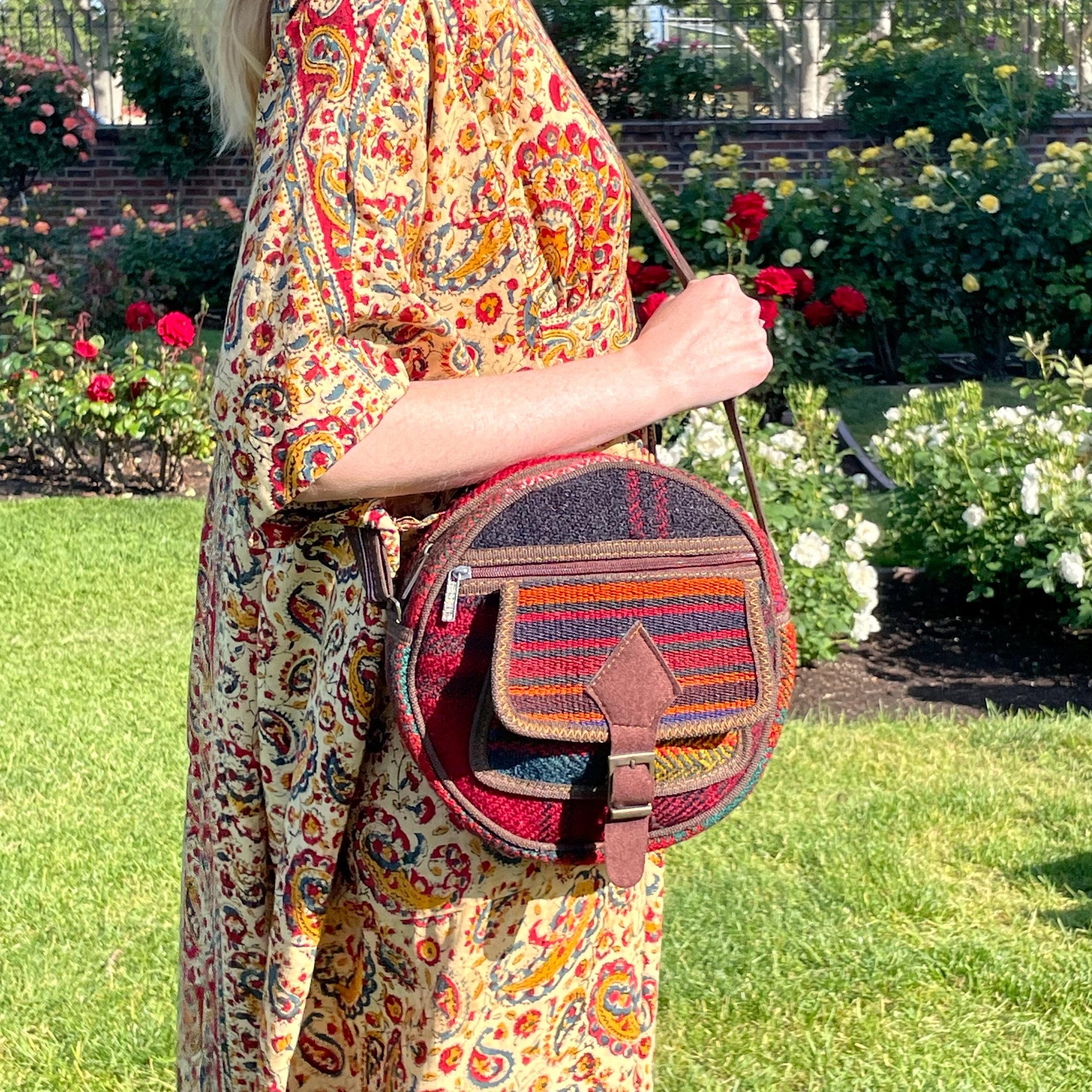 view from the front of a colorful circle bag modeled on a woman in a garden. the purse has handwoven sections of wool, trimmed with brown fabric. The bag has a long strap, front pockets, zippers, and a faux buckle over a snap closure